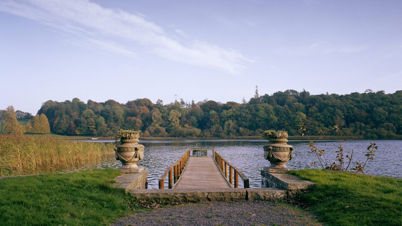 A wooden pier with ornate stone planters on either side extends into a calm lake, with trees lining the far shore under a clear sky.