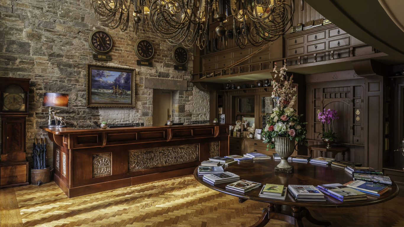 A hotel reception area with a wooden front desk, stone wall, multiple clocks, a large flower arrangement on a round table, and books spread out on the table.