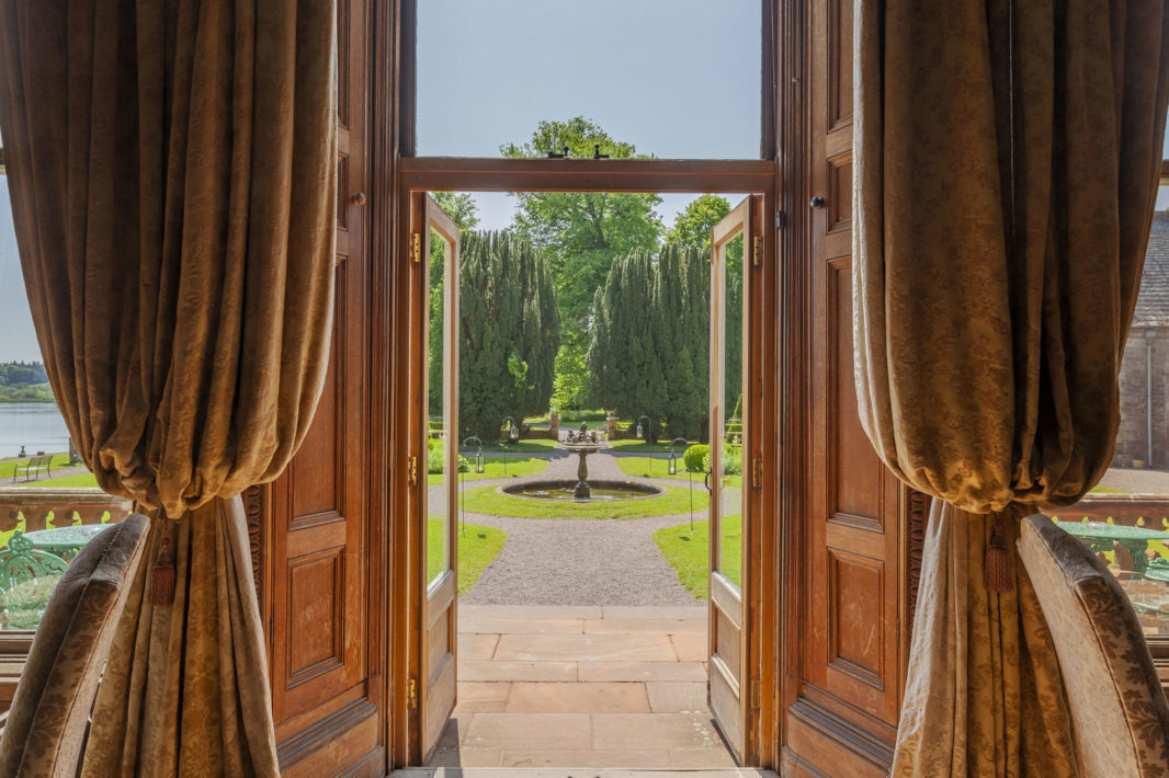 View through open wooden doors to a landscaped garden with a circular fountain, gravel paths, trimmed trees, and green lawns on a sunny day.