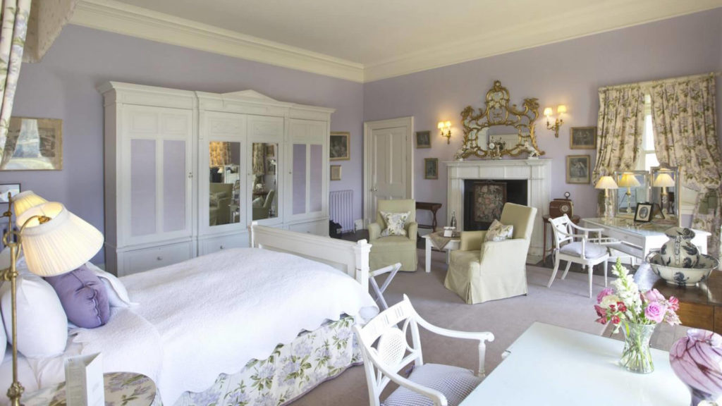 Elegant bedroom with pale purple walls, white furniture, a large bed, ornate mirror above a fireplace, floral curtains, and a vase of flowers on a desk.