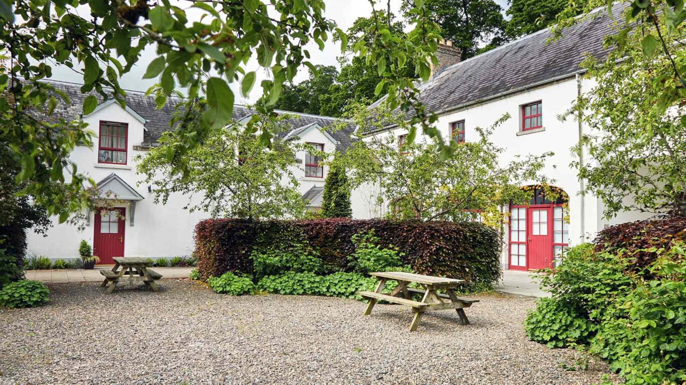 A white two-story building with red doors and windows, surrounded by trees and bushes, features a gravel courtyard with two wooden picnic tables.