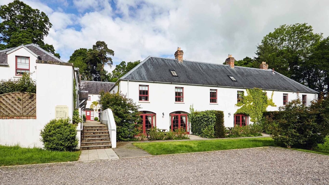 A white two-story house with red-trimmed windows, surrounded by greenery, with a gravel driveway and stone steps leading to the entrance.