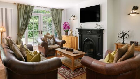 A living room with brown leather sofas, a wooden coffee table, a fireplace with a TV above it, and large windows with green curtains overlooking a garden.