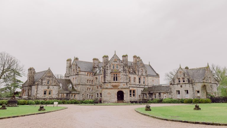 A large, stone mansion with multiple gables, chimneys, and arched entrance, surrounded by a gravel driveway and green lawns under an overcast sky.