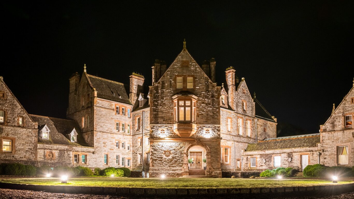A large stone mansion with gabled roofs and lit windows is photographed at night, with exterior lights illuminating the building and surrounding lawn.