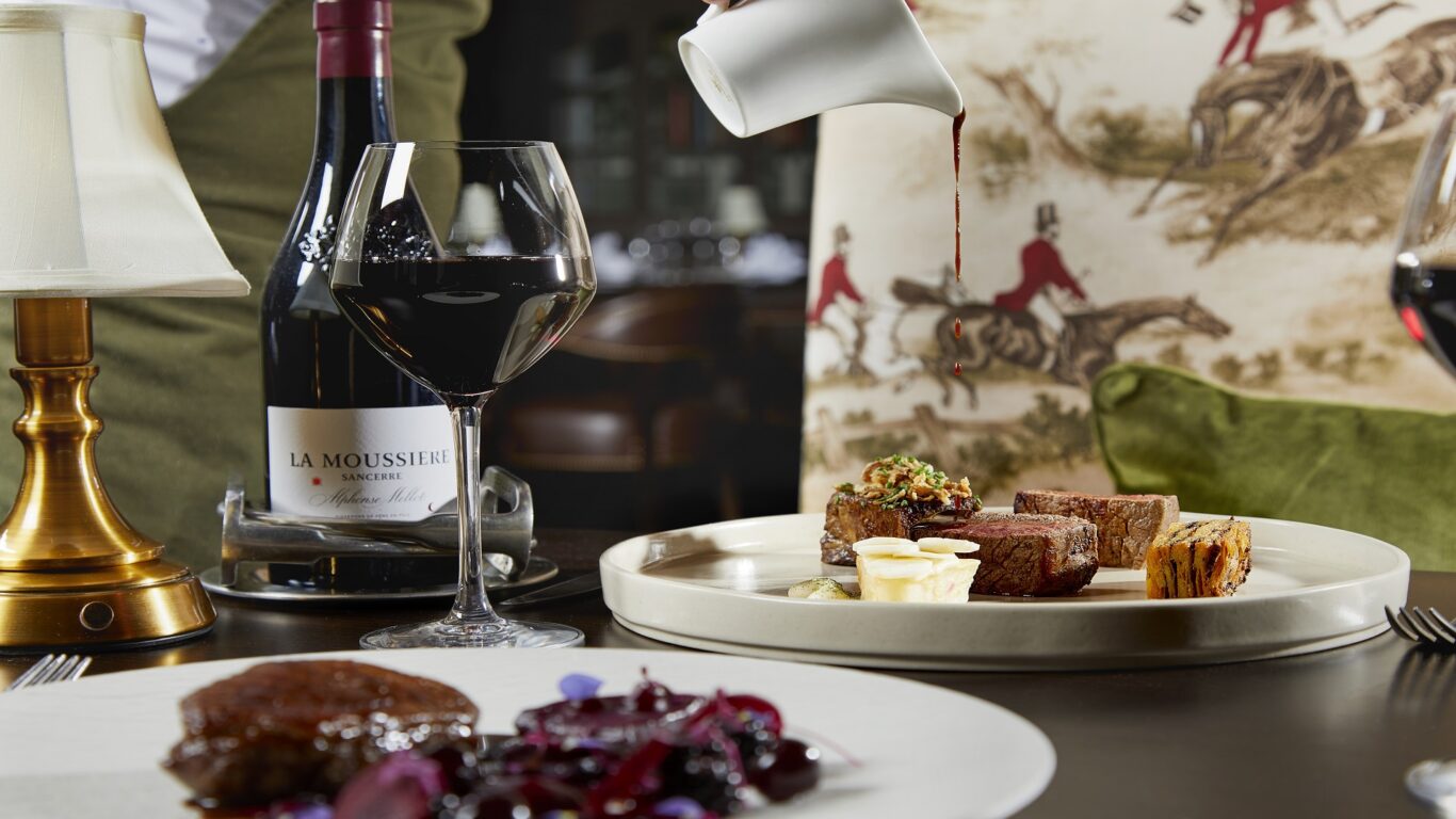 A waiter pours sauce onto a plate of assorted meats and sides, next to a glass and bottle of red wine on a fine dining table.