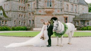 A bride and groom kiss in front of a large stone building, holding a bouquet, with a white horse wearing a green garland standing beside them.