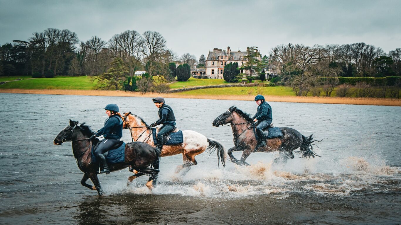 Three people ride horses through shallow water in front of a large historic building surrounded by trees and grassy lawns under a cloudy sky.