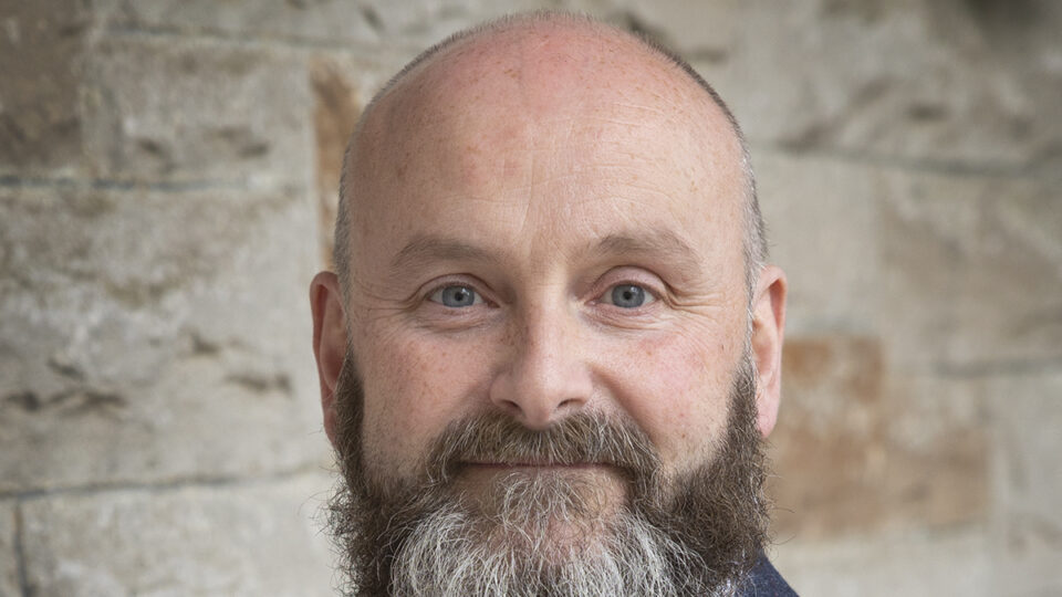 A middle-aged man with a beard, wearing a white shirt and dark blazer, stands in front of a stone wall, facing the camera.