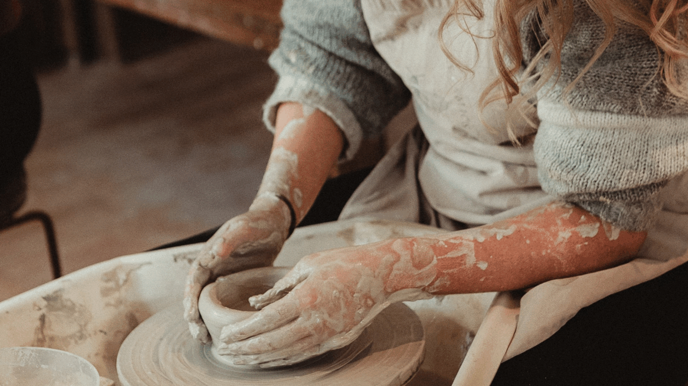 Person shaping clay on a pottery wheel, wearing a light-colored apron and sweater, with hands covered in wet clay.
