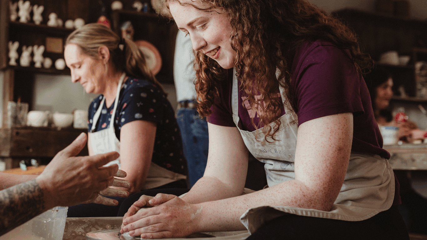 A woman with curly hair smiles while shaping clay on a pottery wheel; others work on pottery in the background.