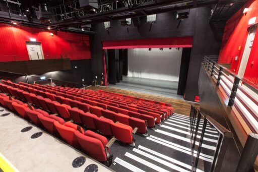 Empty theater auditorium with red seats facing a stage with closed curtains; walls are red, lighting and rigging visible on the ceiling.