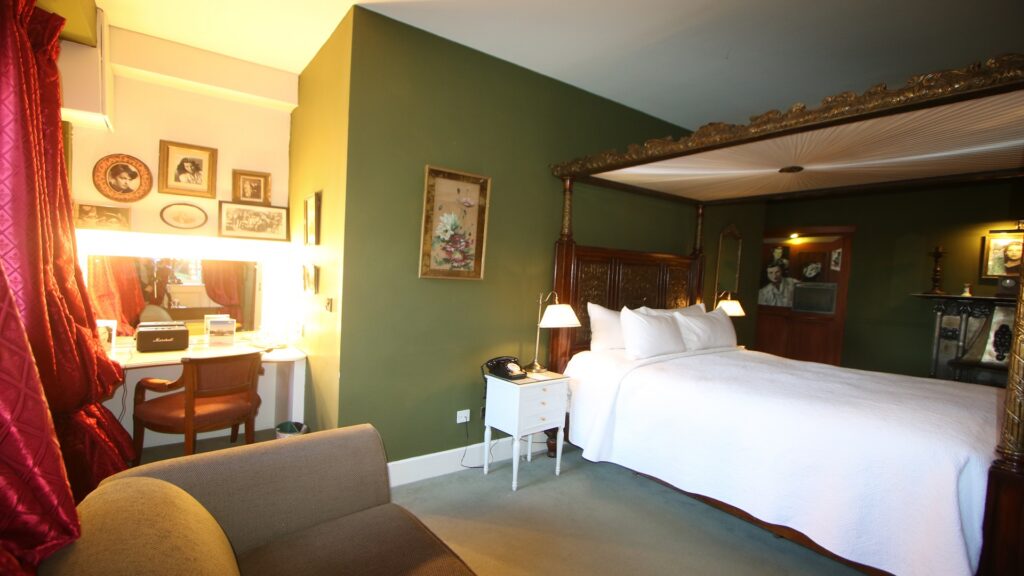 A well-lit hotel room with a four-poster bed, white bedding, a green accent wall, vintage decor, a vanity desk with a mirror, and a brown sofa.