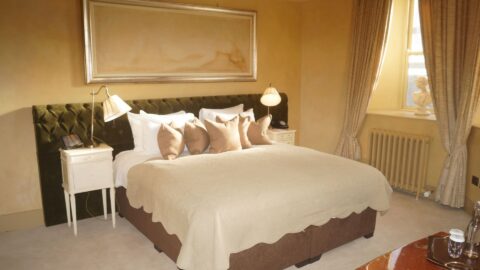 A neatly made double bed with beige bedding and multiple pillows is flanked by two white nightstands with lamps in a warmly lit bedroom.