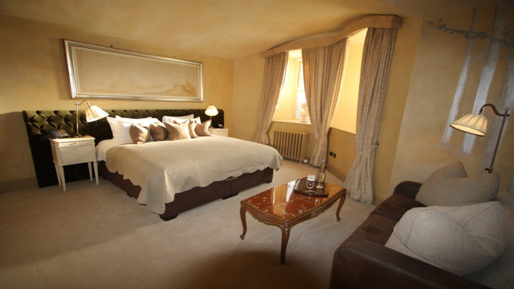 A neatly arranged hotel room with a double bed, nightstands, lamps, a small table with bottled water, a sofa, and tall curtains covering a window.