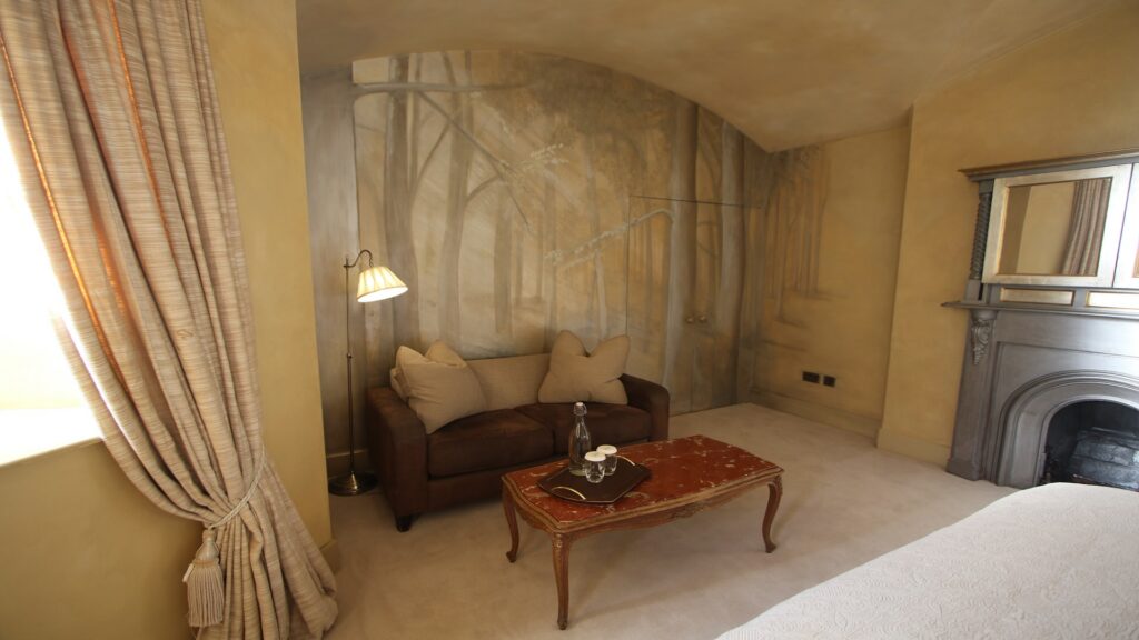 A small living area with a brown sofa, two beige pillows, a tray with a glass and bottle on a brown coffee table, and a wall mural of trees behind the sofa.