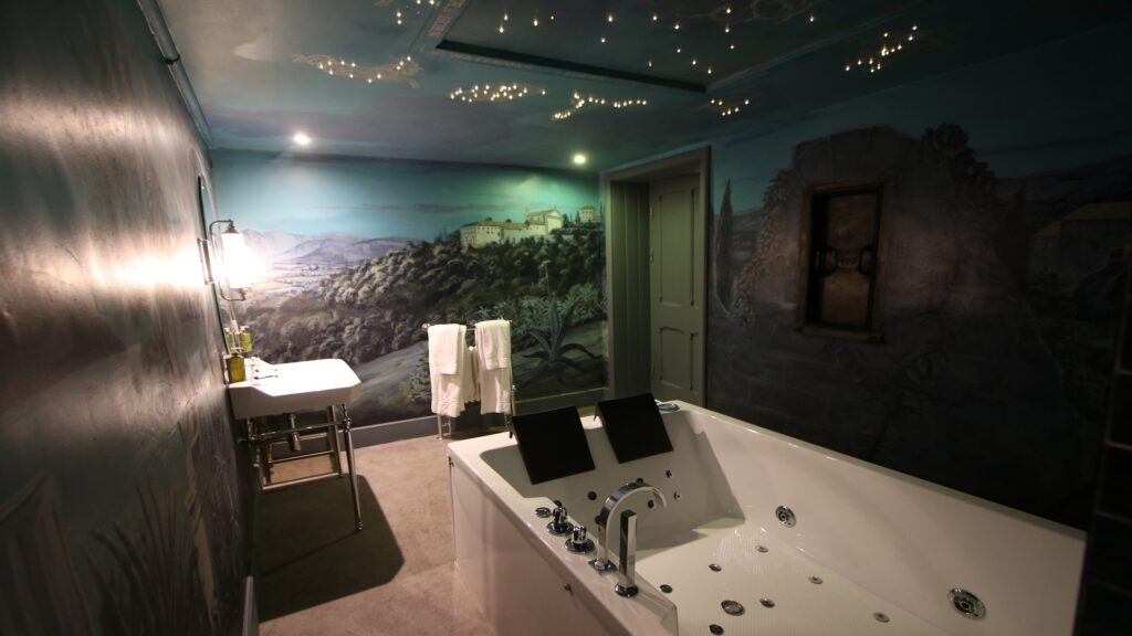 A modern bathroom with a large jacuzzi tub, a wall-mounted sink, towels, and mural-style wall art depicting a landscape with mountains and buildings.