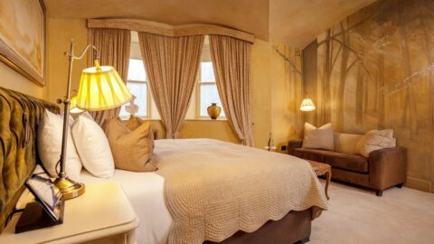 Cozy bedroom with a large bed, neutral bedding, two windows with curtains, a small couch, warm lighting, and a mural of trees on the wall.