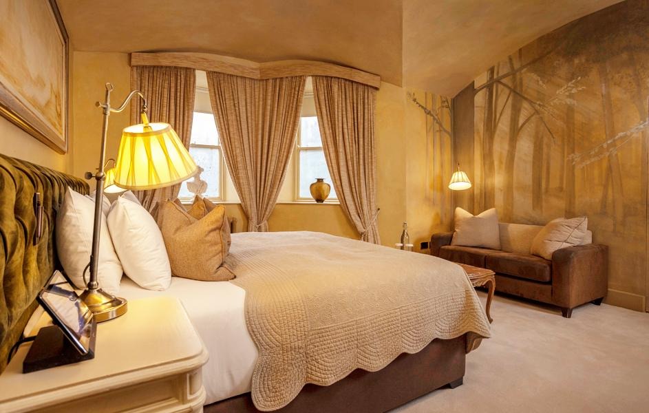 Cozy bedroom with a large bed, neutral bedding, two windows with curtains, a small couch, warm lighting, and a mural of trees on the wall.