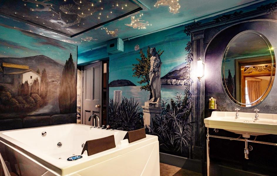 A luxurious bathroom with a large white jacuzzi tub, a double sink, an oval mirror, and walls painted with scenic murals of landscapes and statues.
