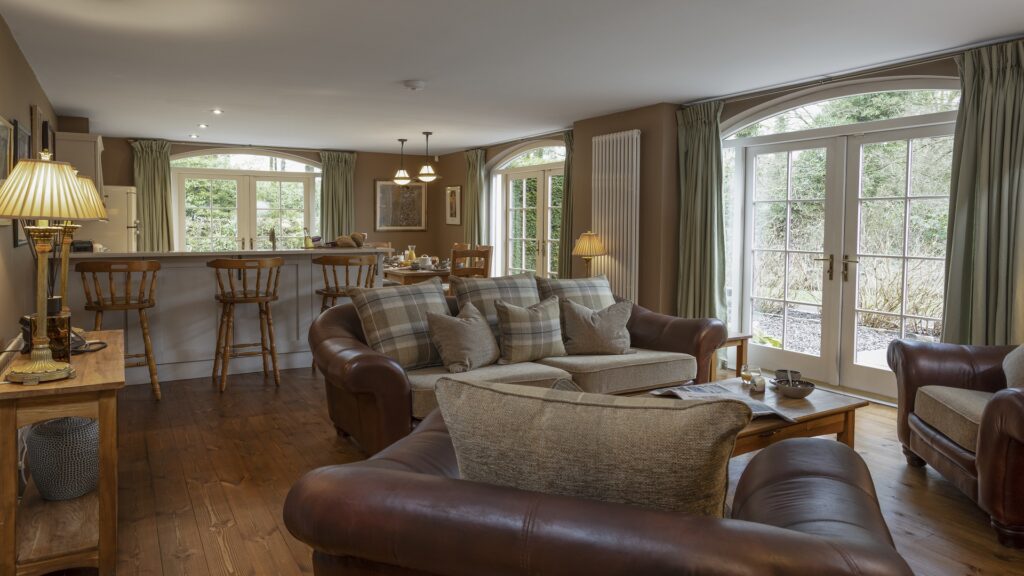CASTLE LESLIE INTERIORS A cozy living room with brown leather armchairs, a plaid sofa, a wooden floor, and large windows letting in natural light, next to a breakfast bar with stools and a dining area.