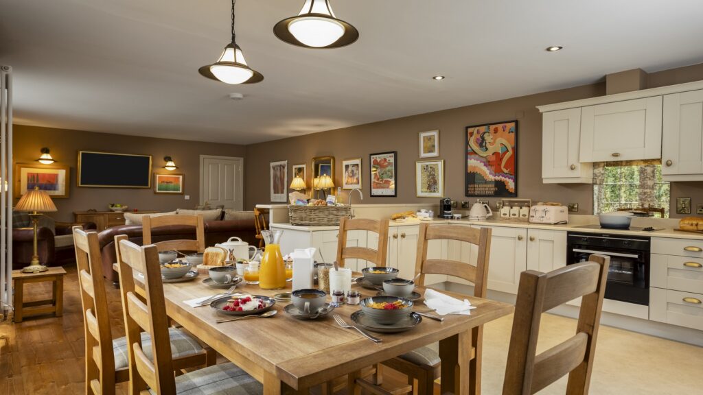 CASTLE LESLIE INTERIORS A kitchen and dining area with a wooden table set for breakfast, surrounded by chairs, modern appliances, and framed artwork on tan walls.