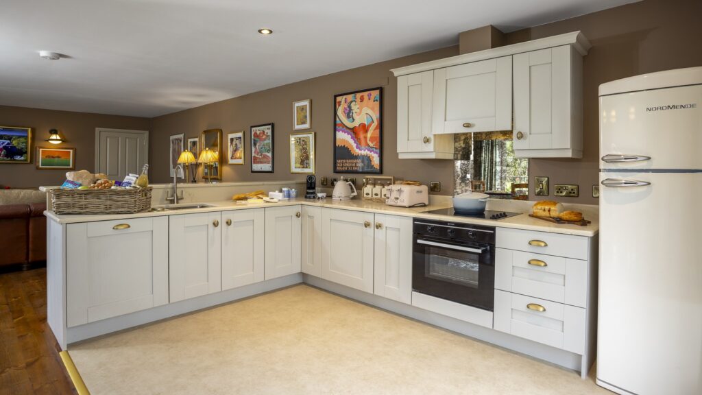 CASTLE LESLIE INTERIORS Modern kitchen with white cabinets, a cream retro fridge, oven, toaster, and kettle. Artwork and framed pictures are displayed on the brown walls. Bread and groceries are on the counter.