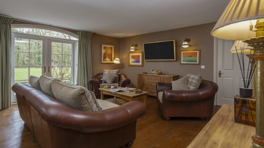 CASTLE LESLIE INTERIORS A cozy living room with two leather armchairs, a sofa, a coffee table, TV, framed artwork, and large windows overlooking a garden.