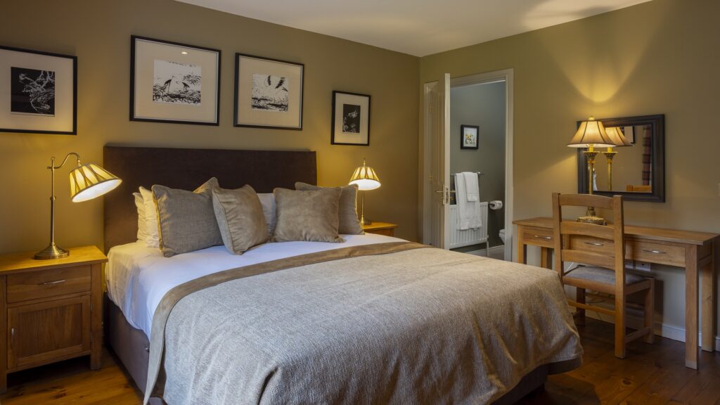 CASTLE LESLIE INTERIORS A neatly made double bed with beige bedding, two nightstands with lamps, framed art on the wall, a wooden desk with a lamp, and an open door leading to a bathroom.