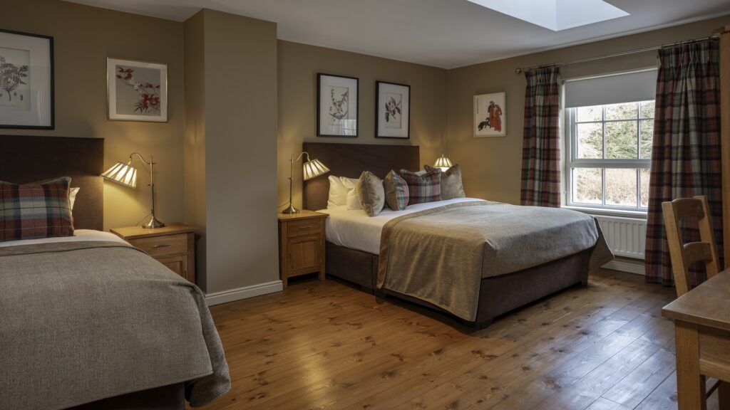 CASTLE LESLIE INTERIORS A cozy bedroom with two beds, wooden floors, plaid curtains, a window with outdoor view, bedside tables with lamps, and framed art on beige walls.