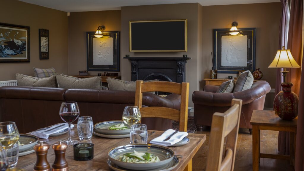 CASTLE LESLIE INTERIORS A cozy living room and dining area with brown sofas, a fireplace, a wall-mounted TV, a dining table set for two, and warm lighting.