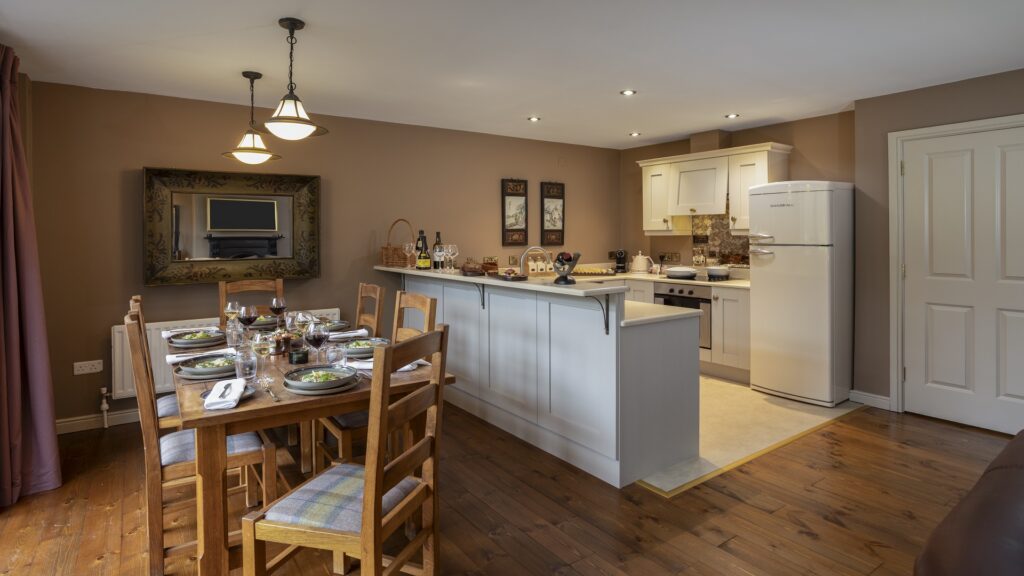 CASTLE LESLIE INTERIORS A dining area with a set table for six, next to a kitchen with white cabinets, a refrigerator, oven, and decorative items on the countertop.