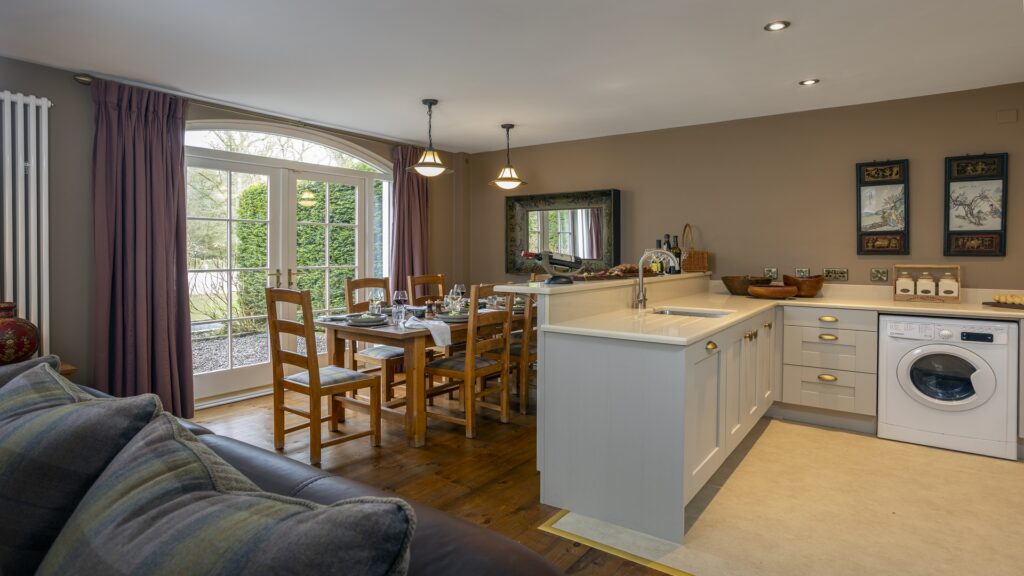 CASTLE LESLIE INTERIORS Open-plan kitchen and dining area with wooden table, chairs, white cabinets, washing machine, and large glass doors overlooking a garden.