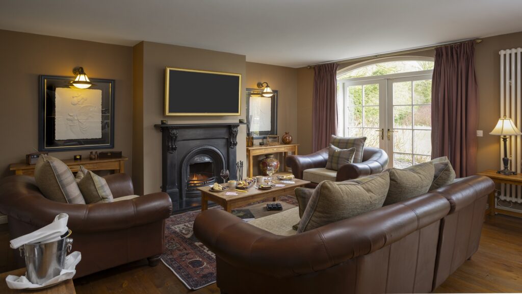 CASTLE LESLIE INTERIORS A living room with brown leather sofas, a black fireplace, a large TV, coffee table set for tea, and large windows with curtains letting in natural light.