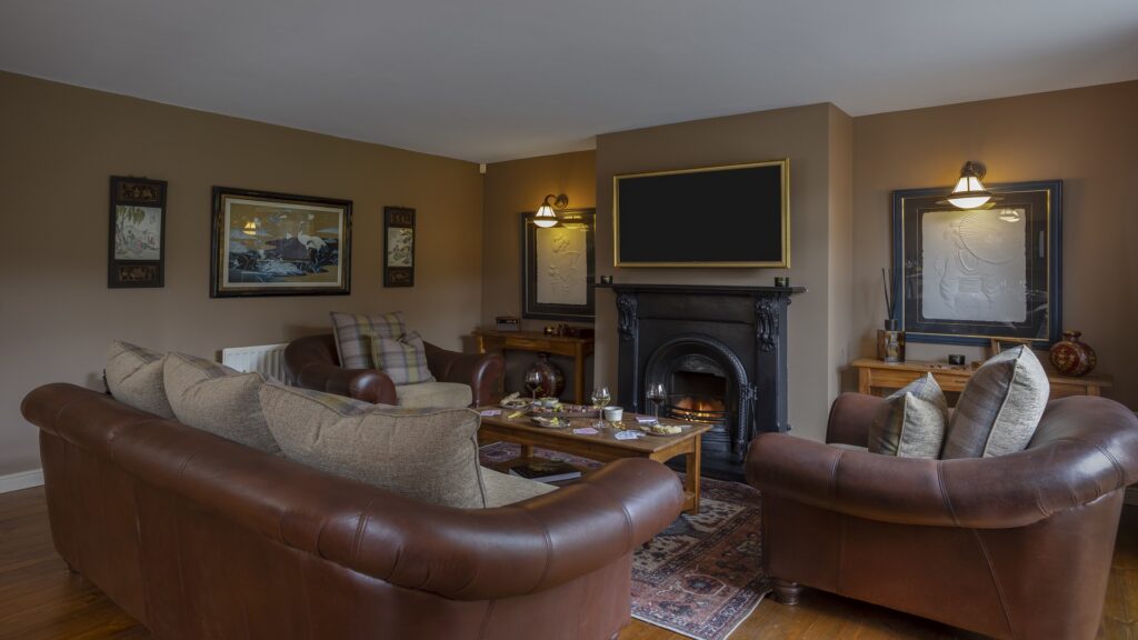 CASTLE LESLIE INTERIORS A cozy living room with brown leather sofas, a dark fireplace, wall art, a large TV above the mantle, and wooden coffee tables with tea items on top.