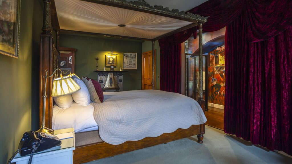 A four-poster bed with a white quilt stands in a room with green walls, framed art, a fireplace, and red velvet curtains.