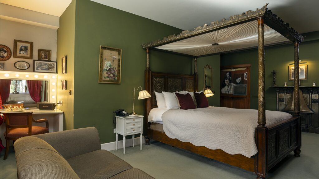 A bedroom with a green accent wall, a large four-poster bed, vintage decor, a sofa, and a vanity table with a lighted mirror.