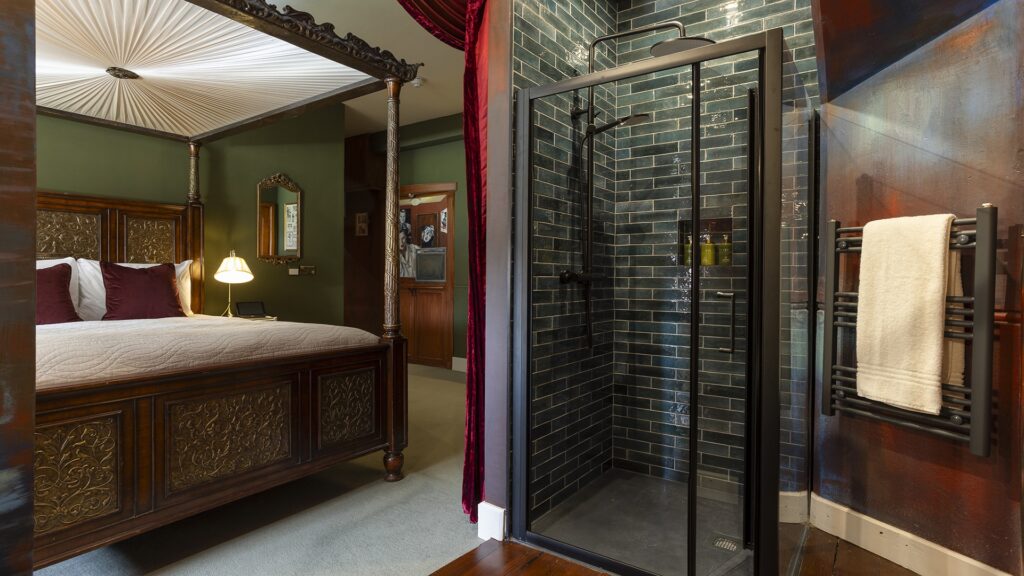 A bedroom with a wooden four-poster bed, a bedside lamp, and an en-suite glass shower next to a heated towel rack holding a white towel.