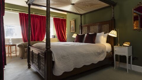 A four-poster bed with white bedding in a green-walled room, red curtains, bedside tables with lamps, an armchair, and a window letting in natural light.