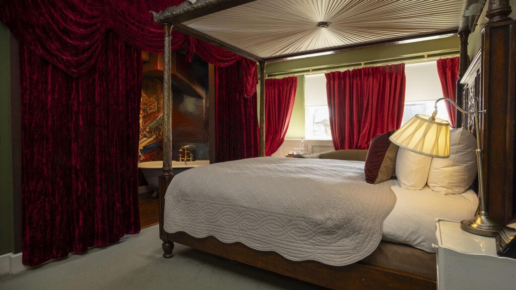 A four-poster bed with white bedding and a canopy sits next to red velvet curtains and a bathtub partially visible in the background.