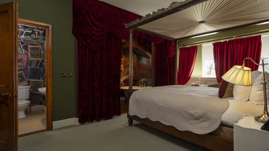 A bedroom with a four-poster bed, white bedding, red velvet curtains, green walls, and an open door leading to a bathroom with posters on the walls.