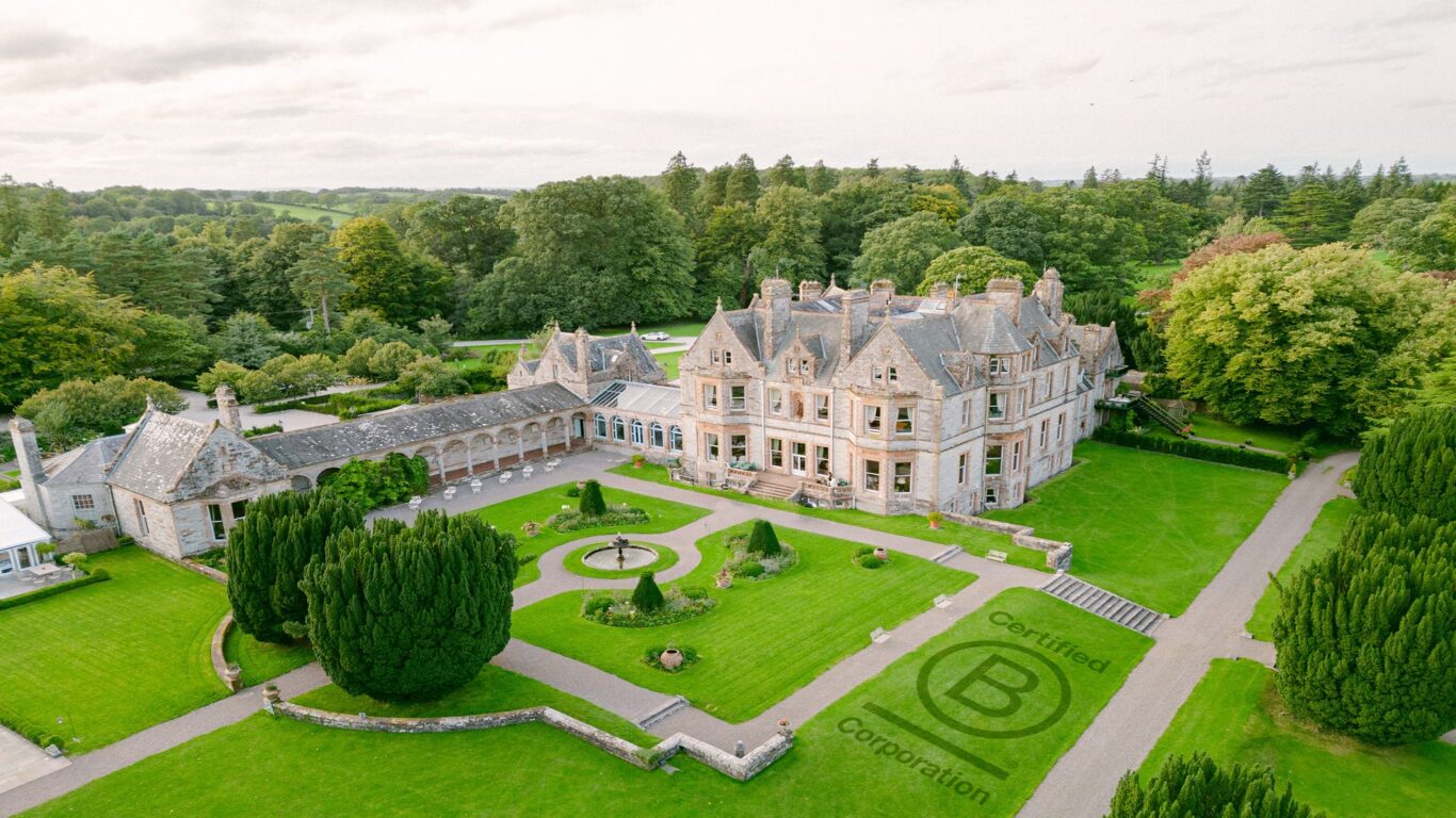 A large historic mansion is surrounded by manicured lawns, gardens, and trees; a 
