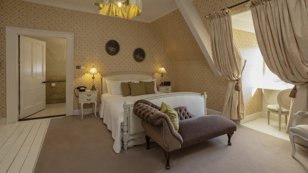 A cozy vintage-style bedroom with a double bed, a brown upholstered bench, two bedside tables, a curtained window, and patterned wallpaper.