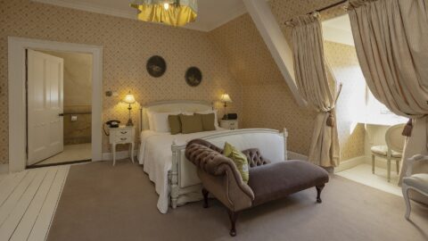 A cozy vintage-style bedroom with a double bed, a brown upholstered bench, two bedside tables, a curtained window, and patterned wallpaper.