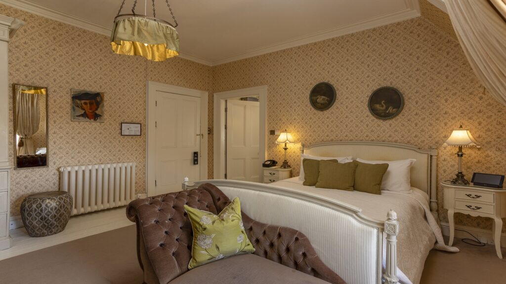 A vintage-style bedroom with patterned wallpaper, a white bed with green pillows, a brown tufted bench, two lamps, and framed art on the walls.