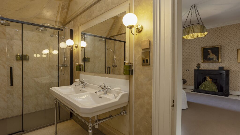 Double sink and mirrored wall in a bathroom with marble-style walls; glass shower enclosure and view into a bedroom with a fireplace and portrait.