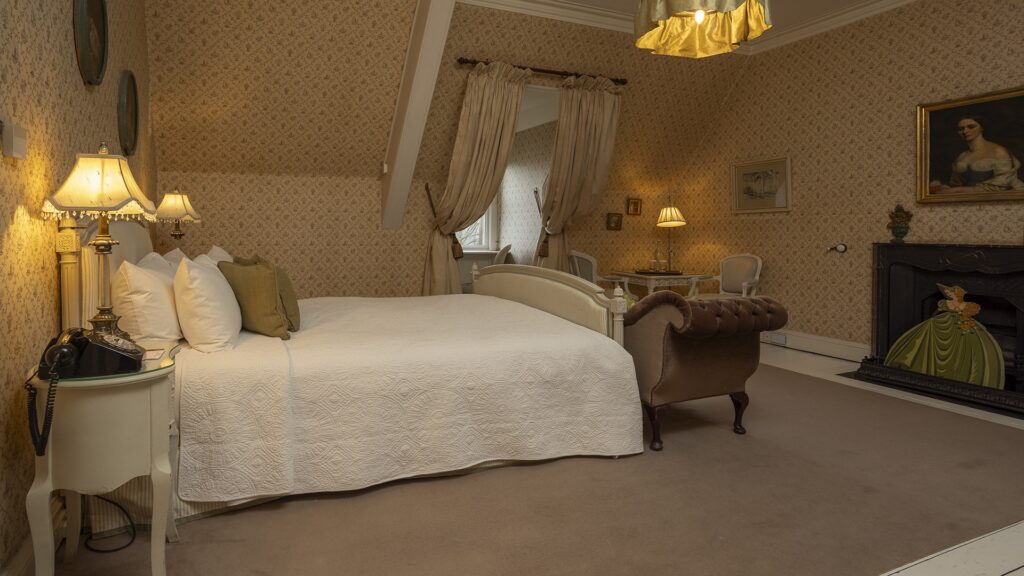 A vintage-style bedroom with patterned wallpaper, a bed with white linens, classic lamps, a brown upholstered bench, a fireplace, and framed portraits on the walls.