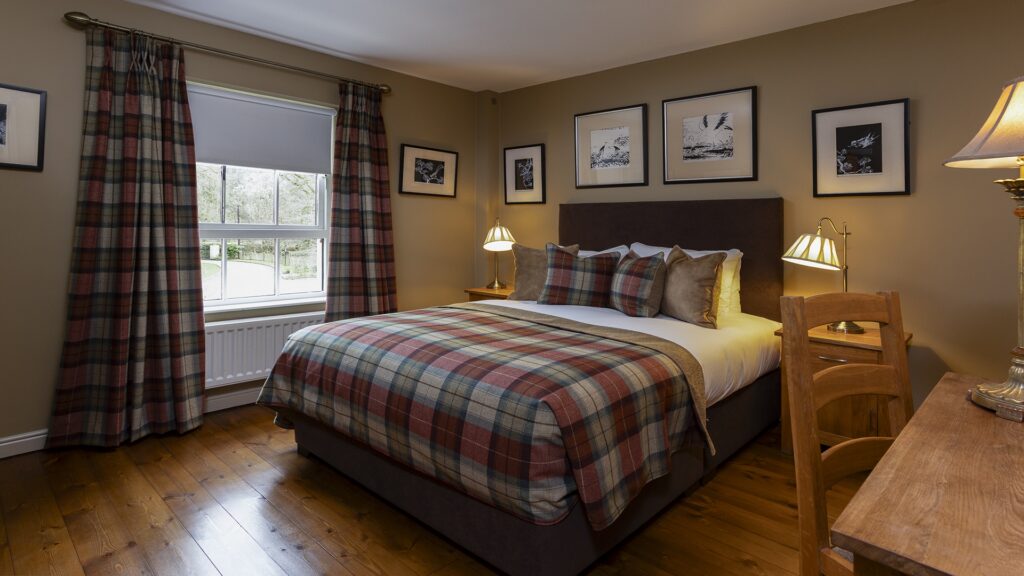A tidy bedroom with a double bed, plaid bedding, wooden floor, matching curtains, framed pictures on the wall, two lamps, and a wooden desk with a chair.