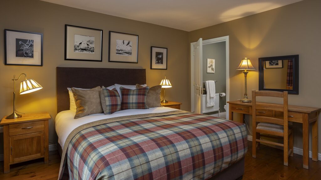 A cozy bedroom with a plaid bedspread, wooden furniture, two bedside lamps, and framed art on beige walls. A door is open near a desk with a mirror, chair, and lamp.