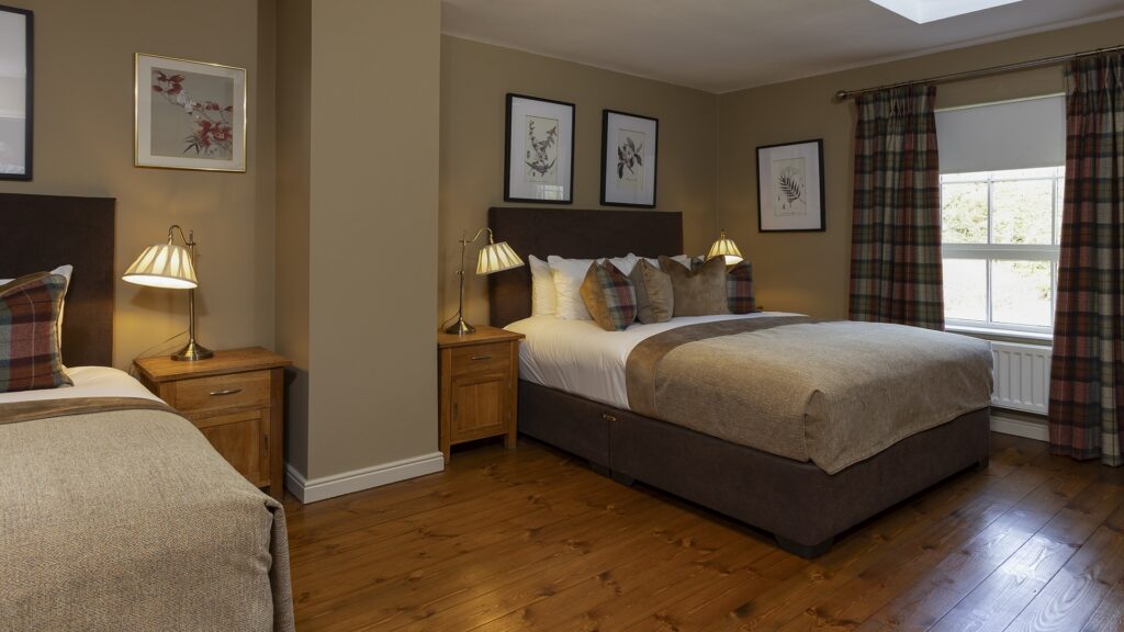 A tidy bedroom with two double beds, plaid pillows, wooden nightstands with lamps, framed art on beige walls, and a window with plaid curtains.